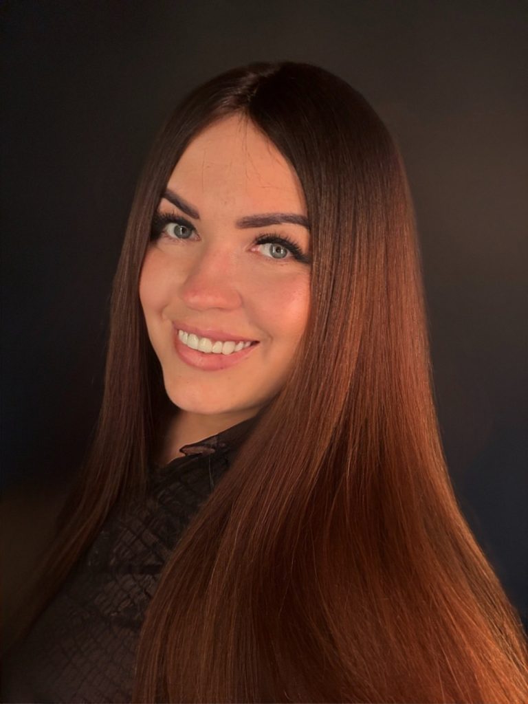 Uśmiechnięty portret kobiety z długimi, brązowymi włosami na ciemnym tle.